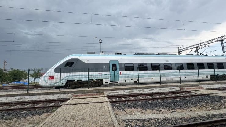 High-speed passenger train in turquoise and white livery at a platform, Turkish flag on the nose, fenced track area, cloudy sky above