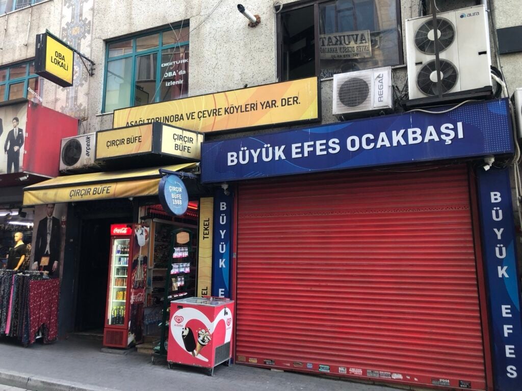 Street view of storefronts with bright yellow ÇIRÇIR BÜFE signs and a blue BÜYÜK EFES OCAKBAŞI sign above a red shuttered shop door on a busy street in Turkey.