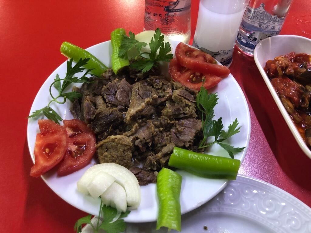 Plate of sliced cooked beef with tomato wedges, onion slices, parsley sprigs, and green peppers on a white plate, set on a red table.