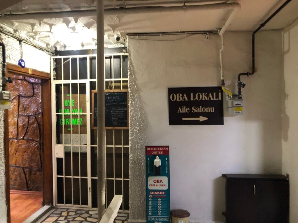 Interior hallway to a cafe, with a black sign reading 'OBA LOKALİ Aile Salonu' and a right arrow, white walls, and exposed pipes.
