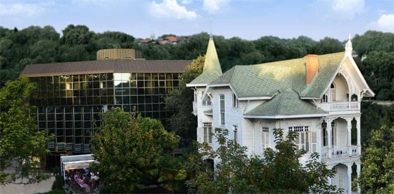 White Victorian-style house beside a glass-front modern building in a green park on a sunny day.