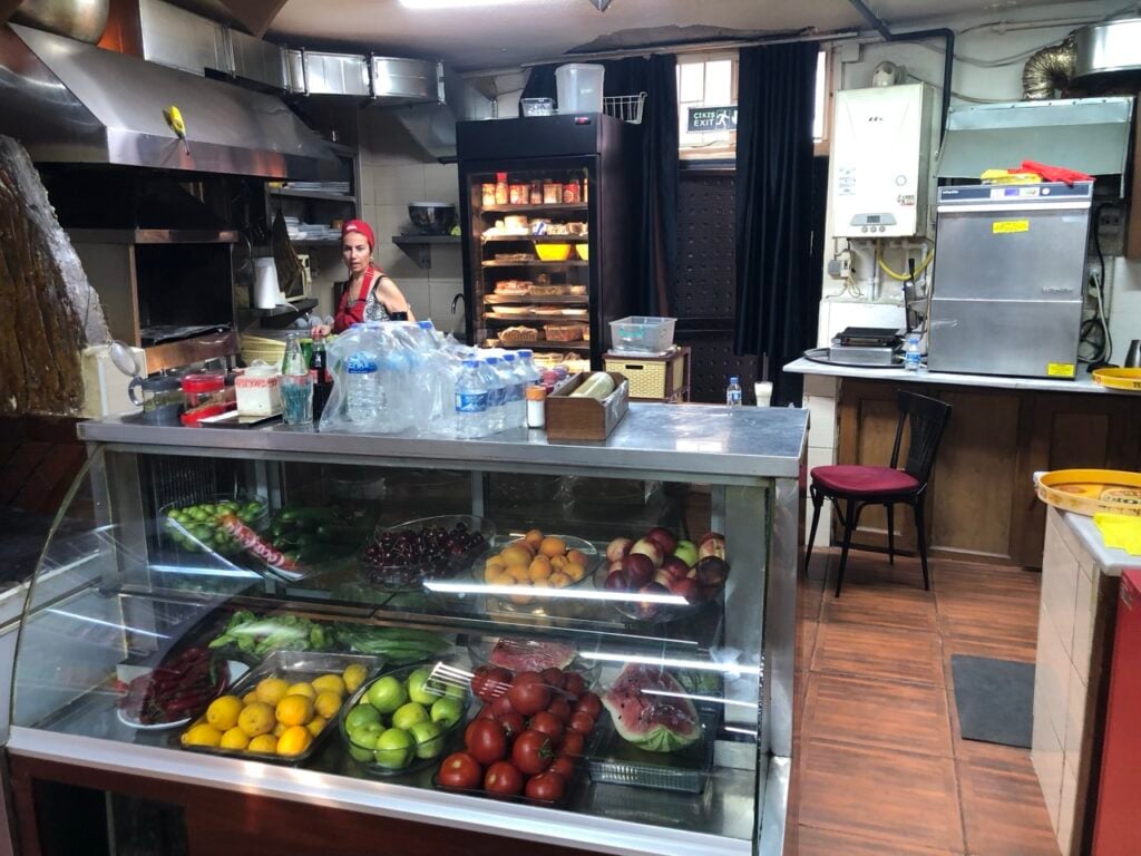 A busy kitchen with a glass fruit display case in the foreground and a female cook in a red bandana behind the counter.
