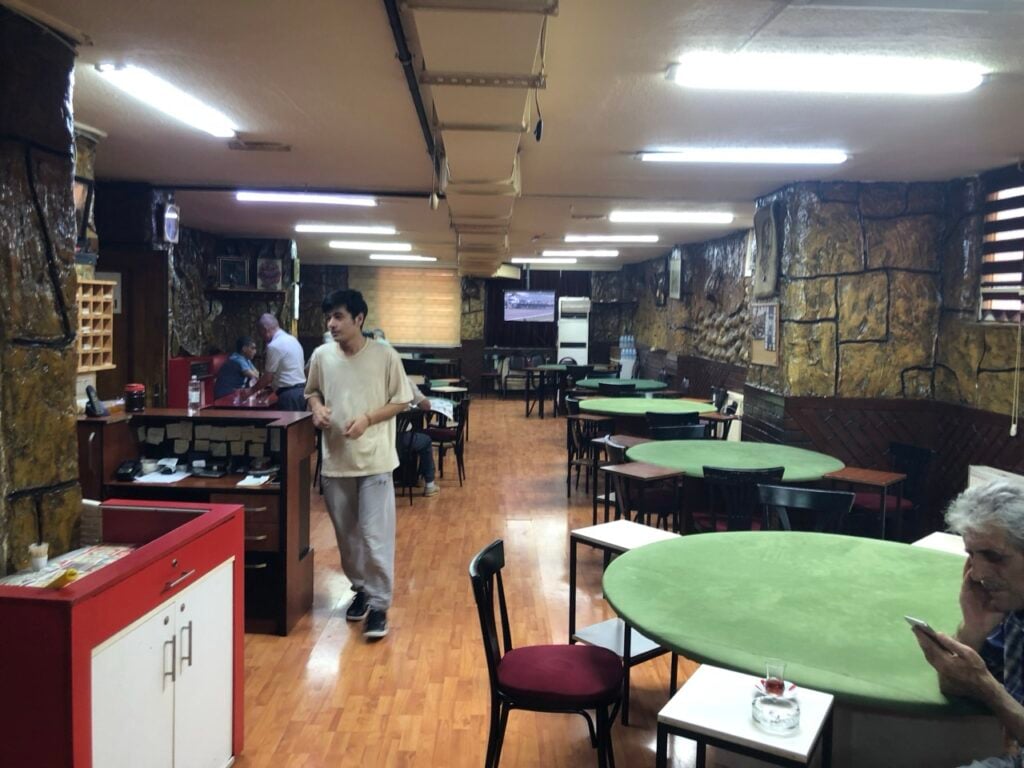 Casual restaurant interior with round green tables, black chairs, and stone walls; a man walks near the counter.