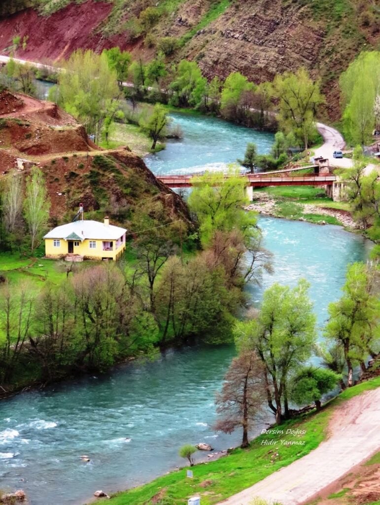 Scenic riverside scene with a turquoise river, a small yellow house, a red bridge, and green trees along a winding road in a hilly landscape.