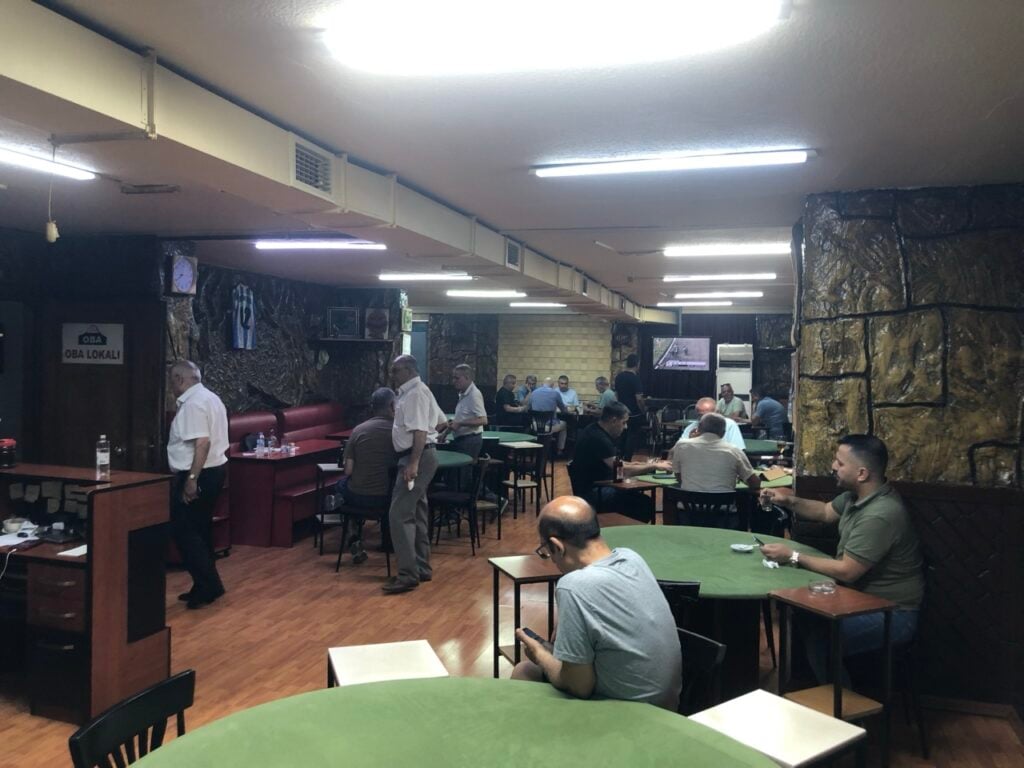 Men seated at round green gaming tables in a stone-walled lounge; fluorescent lights and a TV screen in the background.