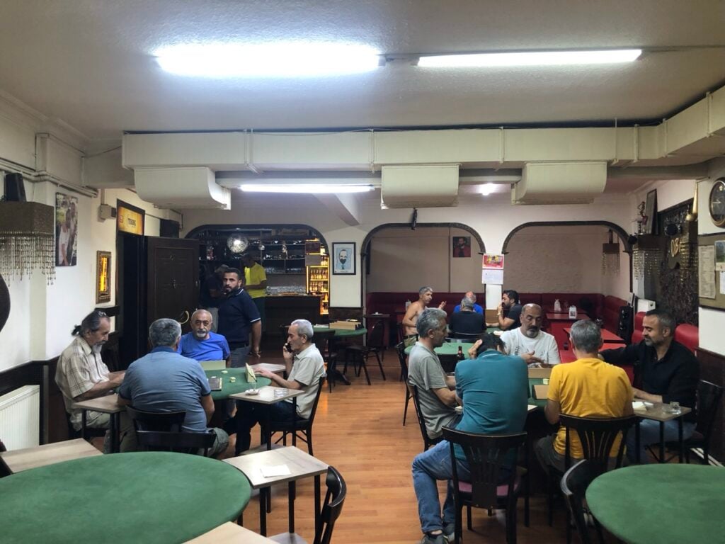 A busy, casual cafe with groups of men sitting at green tables and chatting under bright ceiling lights, wooden floor.