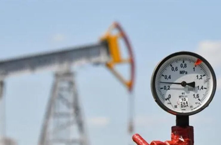 Close-up of a pressure gauge on a red valve attached to a black pipe, with an oil pump blurred in the background.