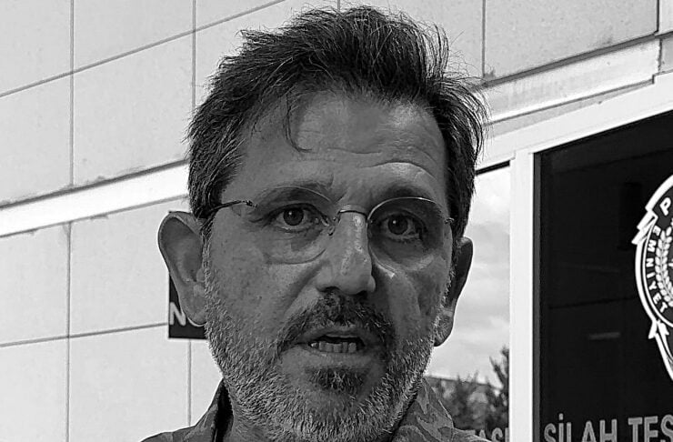 Man with gray hair and beard, wearing glasses and a patterned button-down shirt, outdoors near a building entrance.