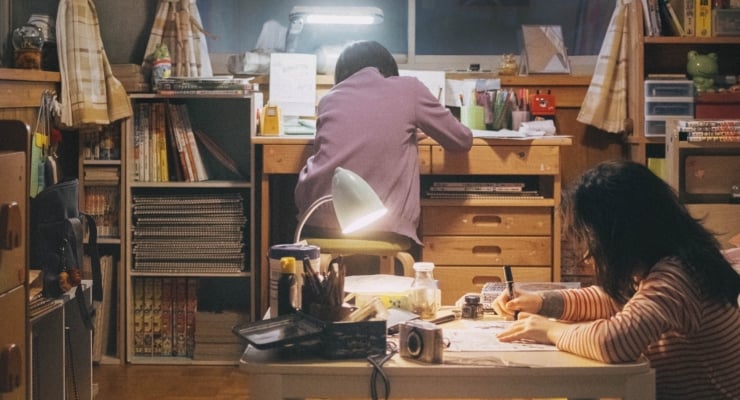 Two people study at a cluttered desk at night; one in a pink hoodie faces away while another writes at the edge of the table under a warm lamp.