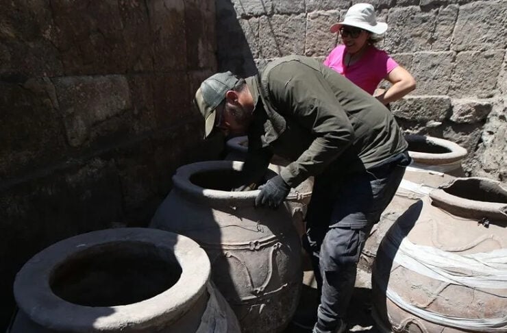 Two people inspect large clay pots in an outdoor, stone-walled area, with one leaning into a pot and the other watching.