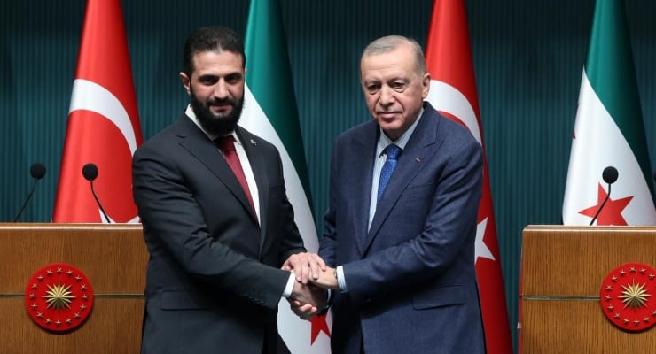 Two men in suits shake hands at a diplomatic event, with Turkish flags and other national flags in the background.