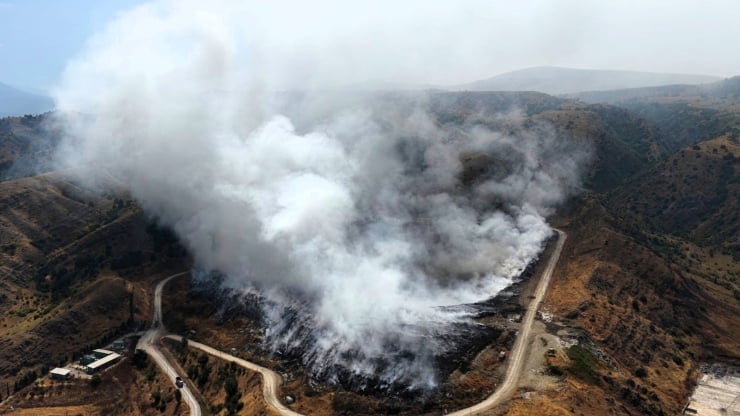 Winding dirt road through hills as a large wildfire emits thick white smoke across the valley.