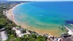 Aerial view of a busy crescent beach with turquoise water, sunbathers, and a parking lot along the shore.