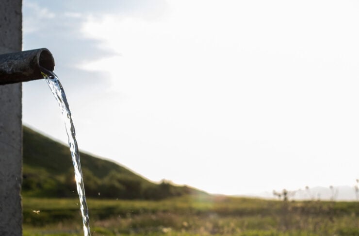 Water flows from a spout into a shallow pool outdoors, with grassy fields and hills in the background at sunset/sun glare on the water surface.