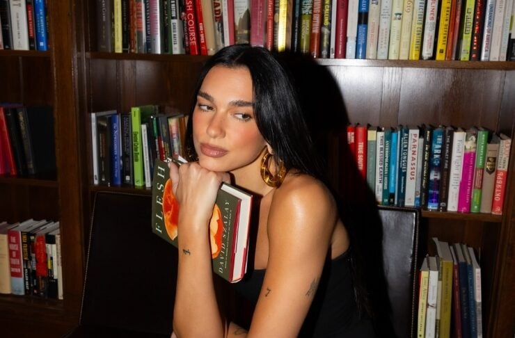 Woman in a black dress and polka-dot tights sits on a leather chair in a library, resting her chin on a book and looking to the side.