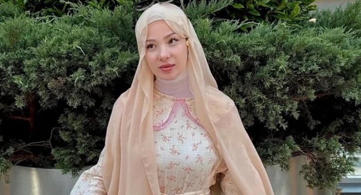 Young woman in a beige hijab and floral dress posing outdoors with green shrubs in the background.