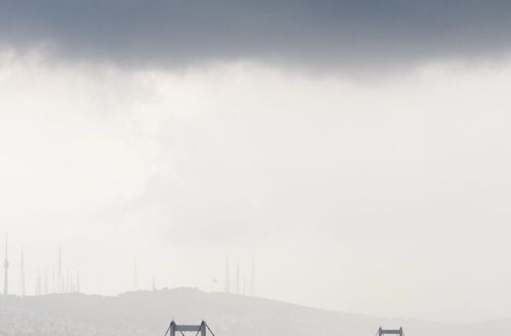 Suspension bridge crossing a river with a city on the shore and misty hills behind under a dark, cloudy sky.