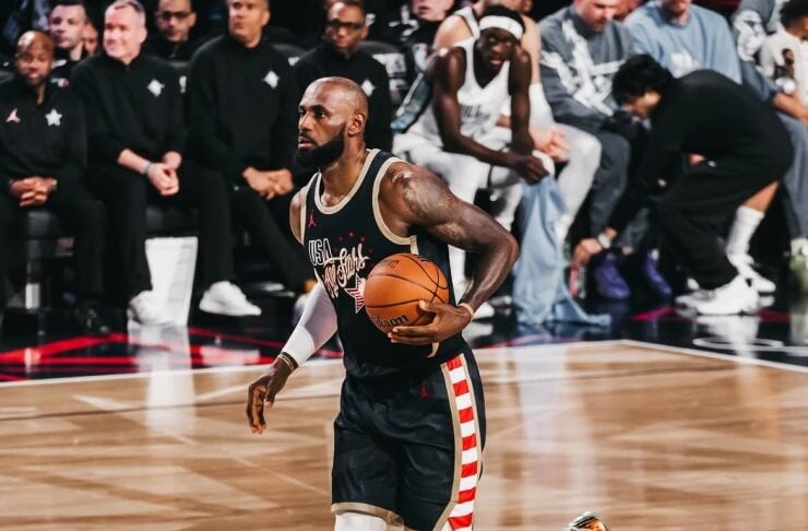 Basketball player in a dark USA-themed jersey dribbles down the court, spectators watching from the bench behind.