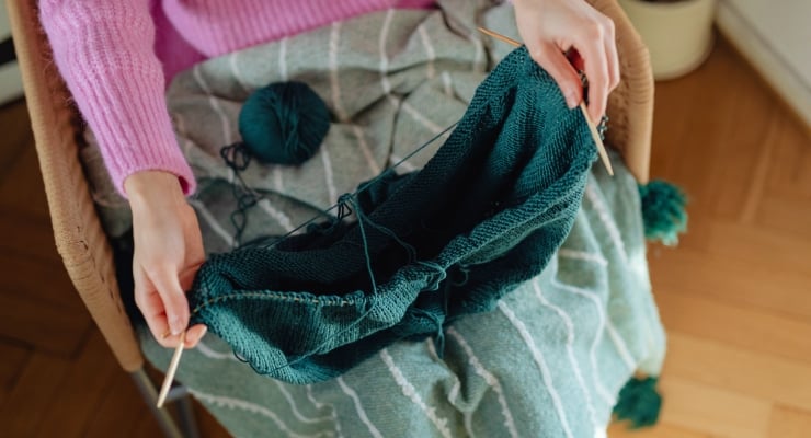 Person knitting teal fabric in a cozy chair, using two knitting needles with yarn nearby and a pink sweater sleeve visible.