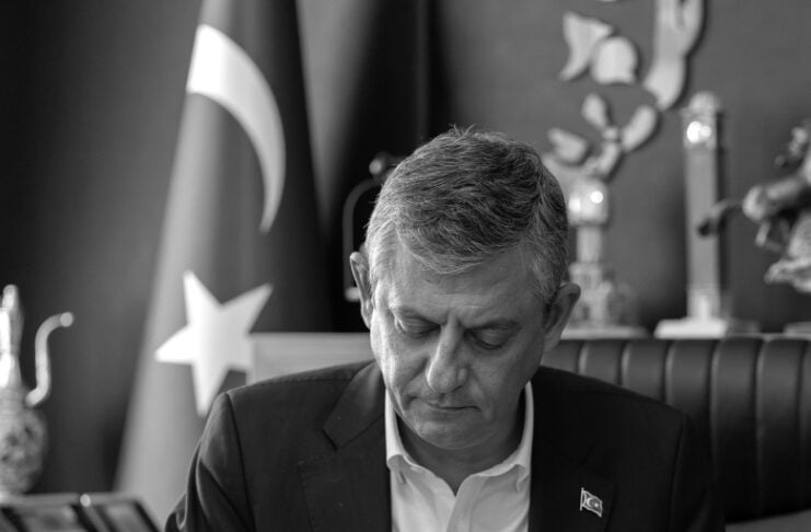 Man in a suit signs a document at a desk in an office, Turkish flag visible in the background.