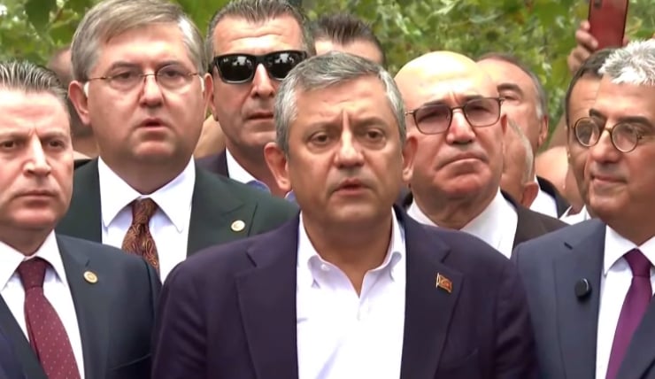 A man in a dark blazer speaks at an outdoor press briefing, surrounded by suited men.