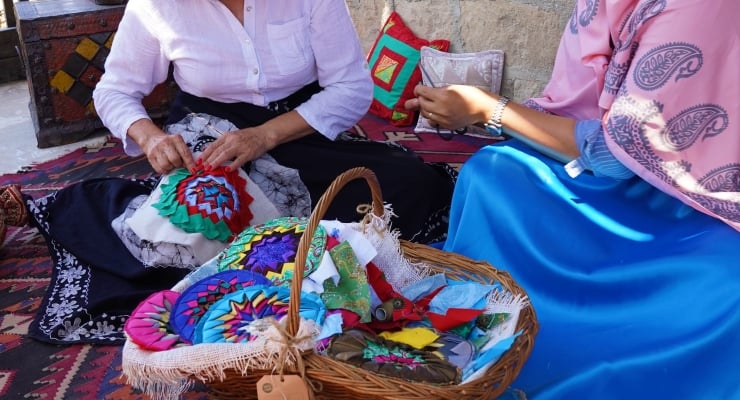 Two people sit on a patterned rug, sewing bright fabric patches as a basket of colorful quilt pieces sits in the foreground.