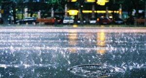 Rain-soaked street with a large pool of water, ripples from heavy rain, and blurred city lights reflected in the wet surface