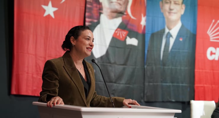 Woman speaks at a white podium with a microphone, olive blazer, in front of red political banners featuring portraits and stars.
