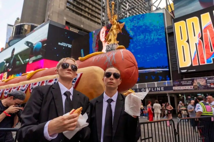 Two men in black suits and sunglasses hold hot dogs in Times Square with a giant pumpkin float behind them and neon billboards nearby.