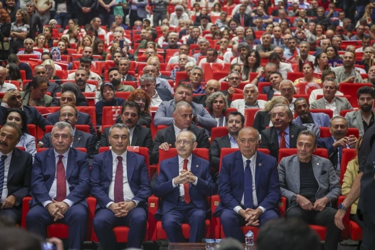 Crowded theater audience with a front row of suited officials in red seats, some clapping, overall attentive to the stage.