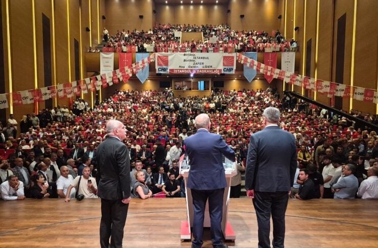 Three men in suits speak from a podium on a stage in a packed auditorium, addressing a large audience with CHP banners overhead.