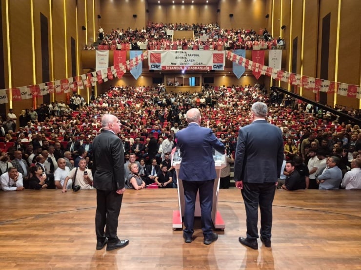 Three men in suits speak from a podium on a stage in a packed auditorium, addressing a large audience with CHP banners overhead.