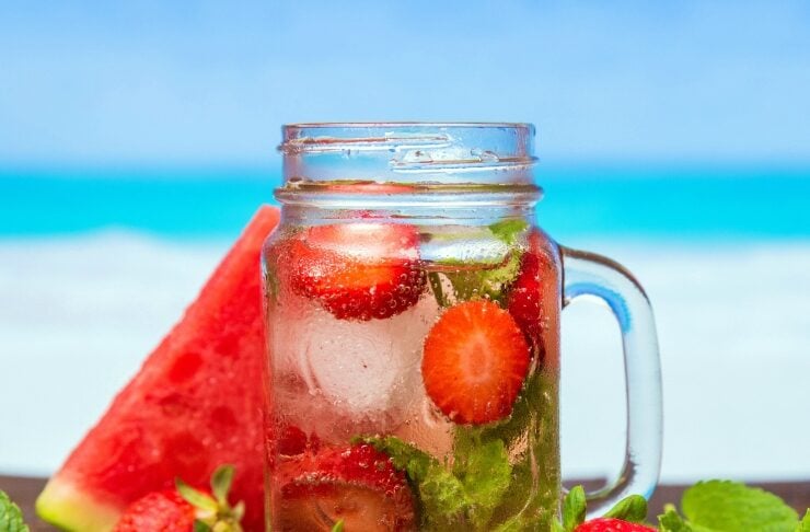 Mason jar with strawberry-mint infused water, surrounded by fresh strawberries and watermelon on a wooden table by the beach.