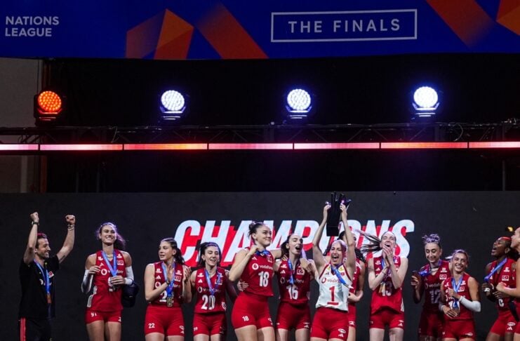 Women’s volleyball team in red uniforms celebrate on stage, raising a trophy with medals against a bold CHAMPIONS backdrop.
