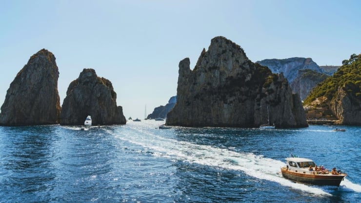 Blue sea with towering rocky sea stacks and a motorboat leaving a white wake across the water.