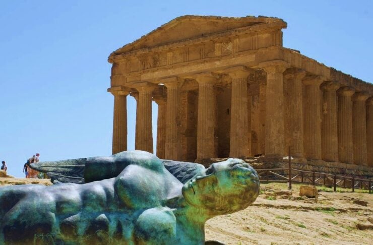 Reclining winged statue in front of an ancient Greek-style temple with tall columns.