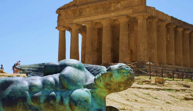 Reclining bronze statue with green patina in the foreground, a columned ancient temple in the background on a sunny day.