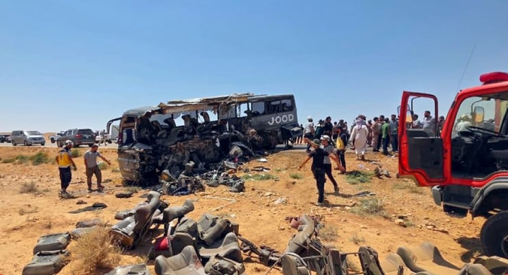 Bus wreck in a desert area with a collapsed bus and scattered debris, people gathering around the scene.