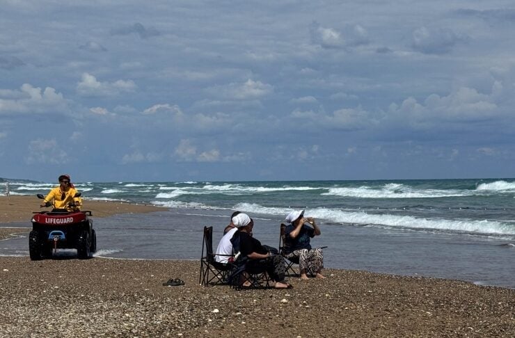Lifeguard on a red ATV parked on a pebbled beach as two people sit in folding chairs near the water, watching the waves under a cloudy sky.