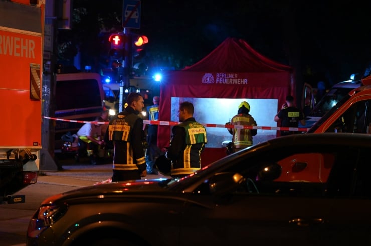 Nighttime emergency scene with firefighters in reflective jackets behind red and white caution tape near a Berliner Feuerwehr tent.