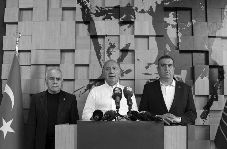 Three men stand at a press conference podium: the center speaker addresses microphones, flanked by two men, with a Turkish flag on the left and a map backdrop behind them.