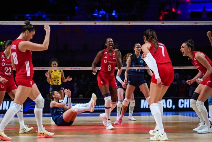 Women’s volleyball team in red celebrates at the net after a rally; one player on the floor gestures with raised arms and teammates cheer nearby.