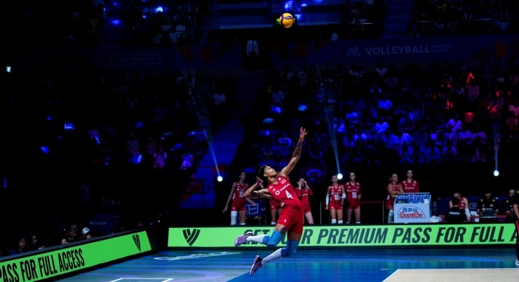 Female volleyball player in red jumps mid-air to spike the ball during a dark arena match, green advertising boards along the sideline behind her.