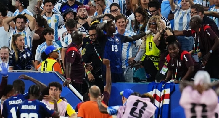 Crowd of fans surrounding players at a bench area, some raising arms in celebration, blue-and-white team jerseys visible.