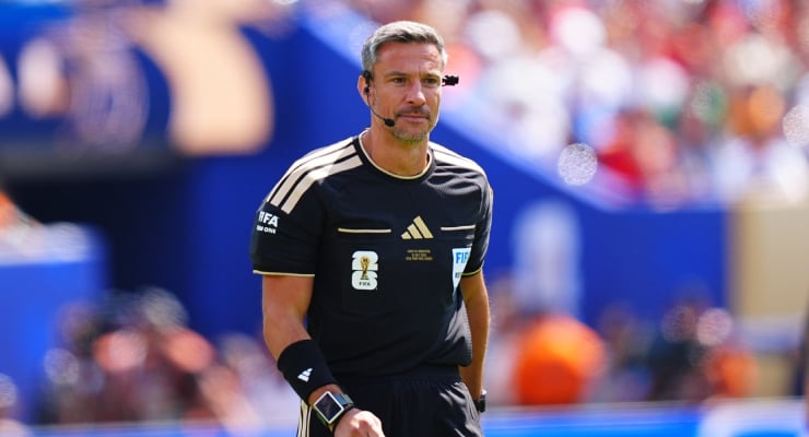 Male soccer referee in a black FIFA uniform with a headset, standing on the pitch during a match.