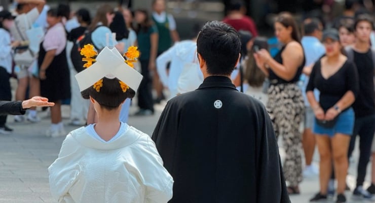 Back view of a graduate in a white gown and square cap walking beside a man in black, with a crowd in the background during a graduation.