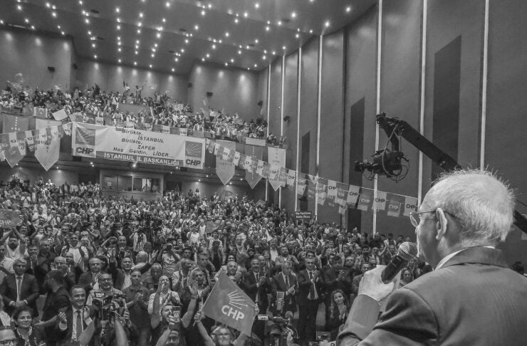 Male politician in a suit speaks into a microphone onstage to a packed indoor rally, crowd waving CHP flags and banners beneath hanging lights.