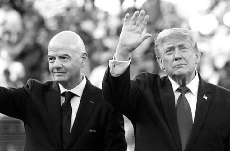 Two men in dark suits wave to a crowd from a stage; the left is bald, the right has light hair and a small flag pin.