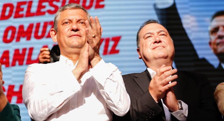 Two men in formal attire clap on a stage at a political rally, with a red and blue backdrop and large Turkish text behind them.