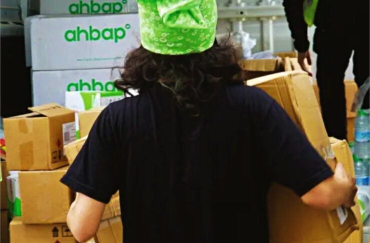 Person seen from behind, wearing a green bandana, carrying a cardboard box in a warehouse with stacked boxes labeled 'ahbap'.